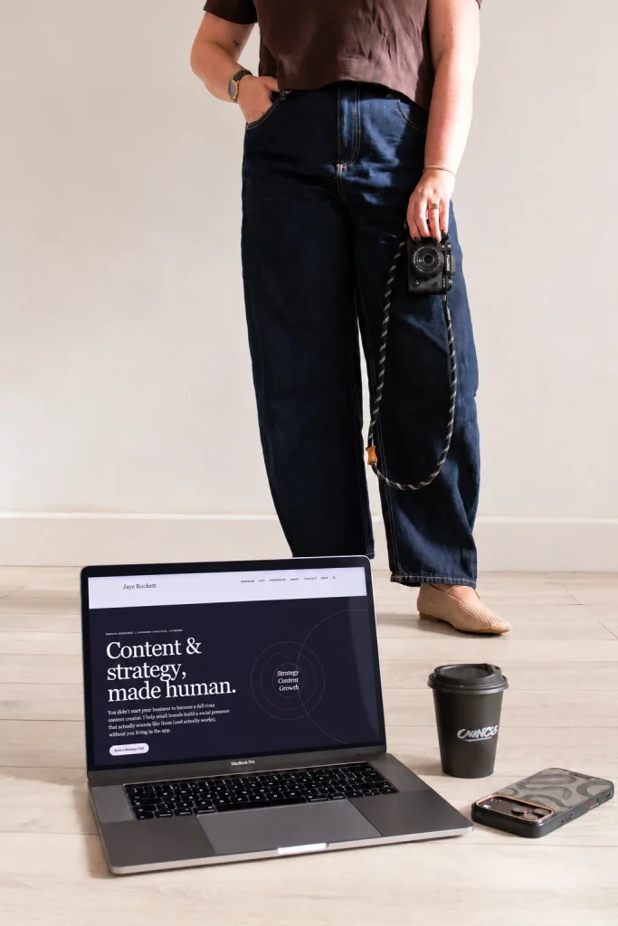 Jaye holding her camera with a laptop, phone and coffee in the foreground