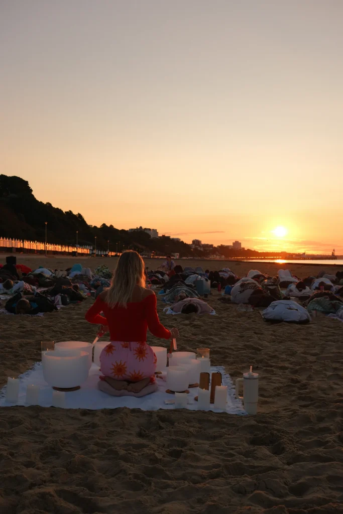 Sound bath facilitator seated with singing bowls as the sun rises over the beach